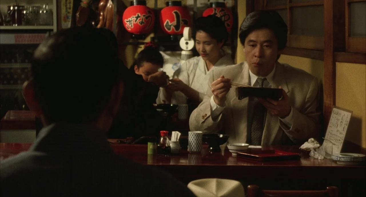 The Discarnates (1988) movie still: medium — An izakaya interior with wooden walls and red lanterns hanging above a counter. A man in a suit eats…