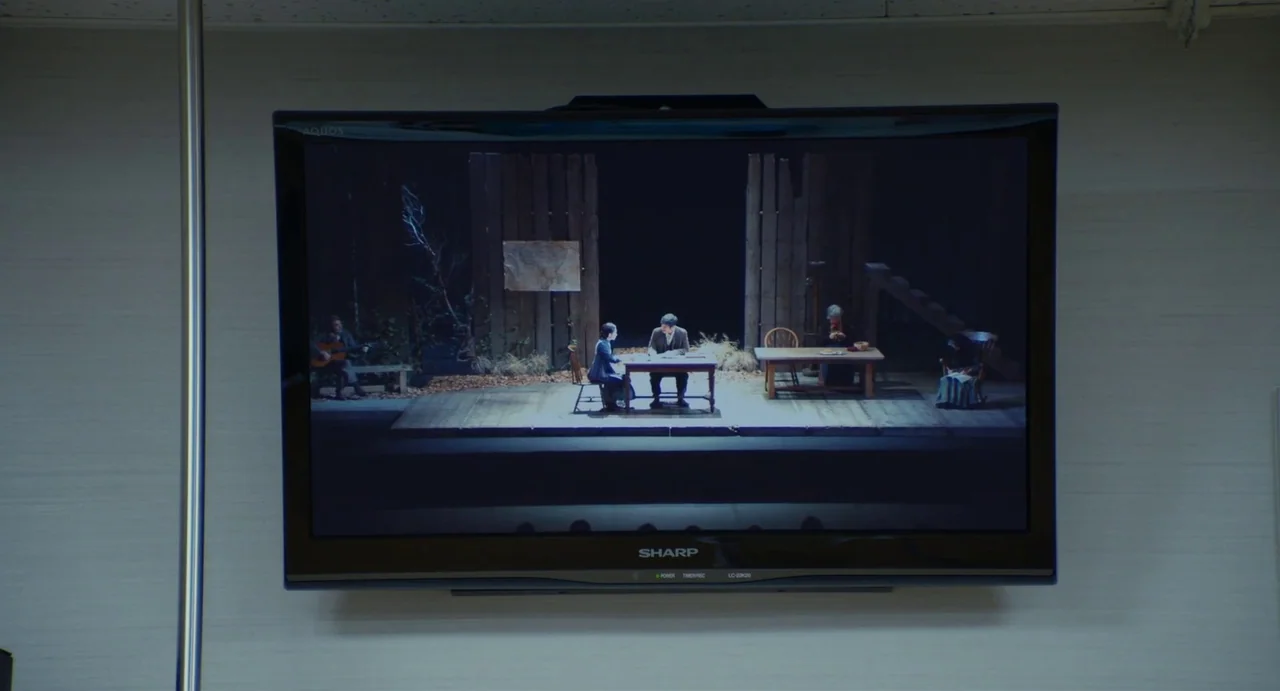 Drive My Car (2021) movie still: wide — A flat-screen television is mounted on a plain, light-colored wall with a vertical metal pole. The screen…