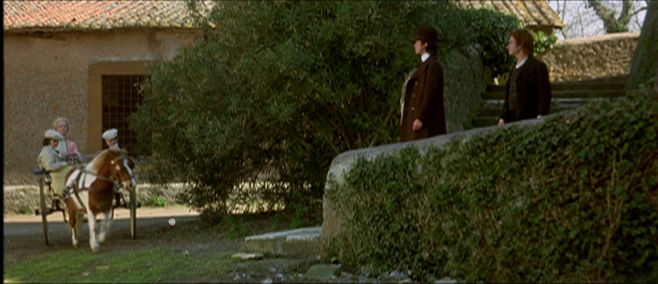 Flesh For Frankenstein (1973) movie still: wide — A stone-walled exterior path leads past thick green bushes toward old rustic buildings. Two men…