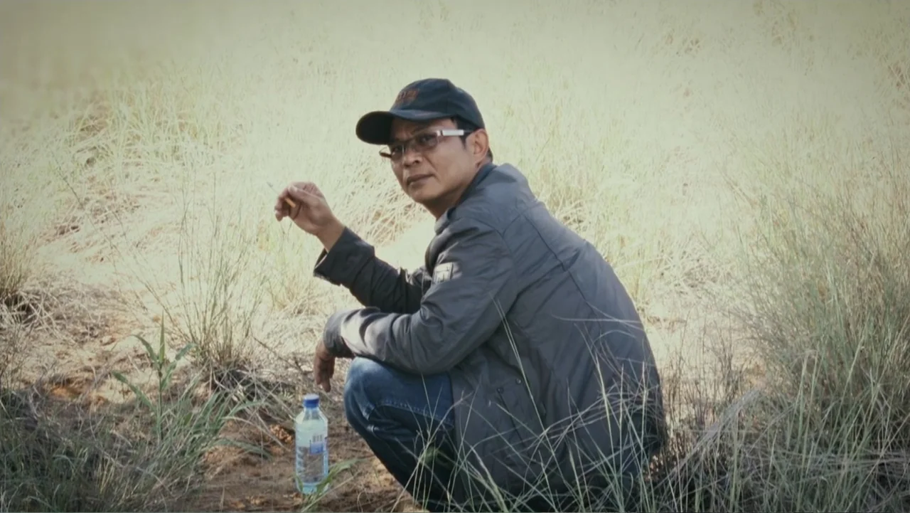 Bloody Trophy (2025) movie still: medium — An open field of tall, dry grass stretches out under bright daylight. A man in a dark jacket squats on…