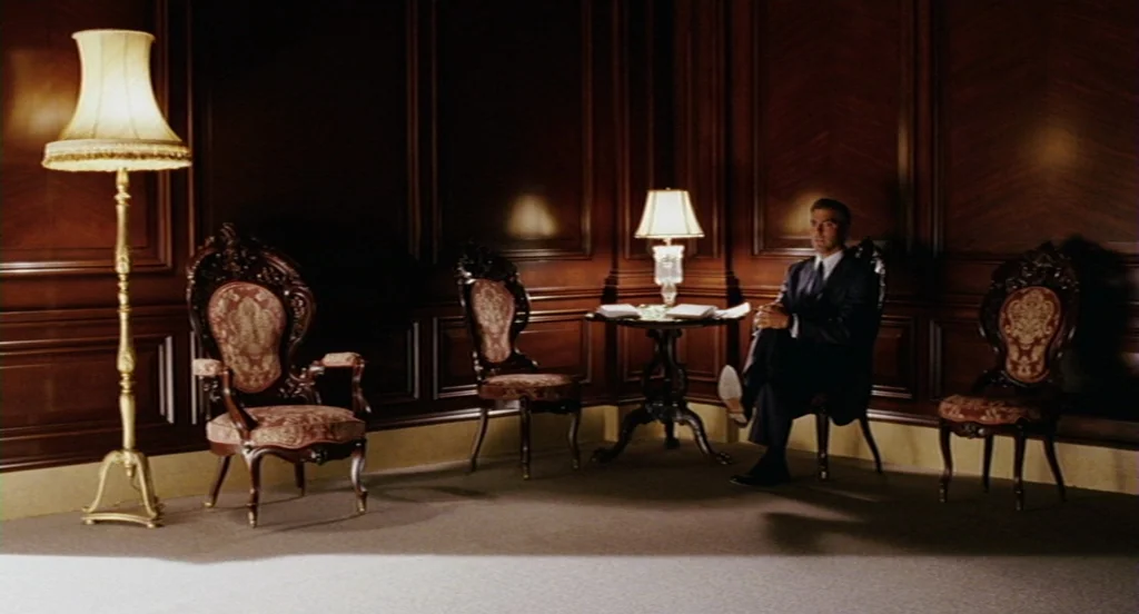 Intolerable Cruelty (2003) movie still: wide — A dark, wood-paneled room is furnished with ornate antique chairs and small tables. A man in a dark…