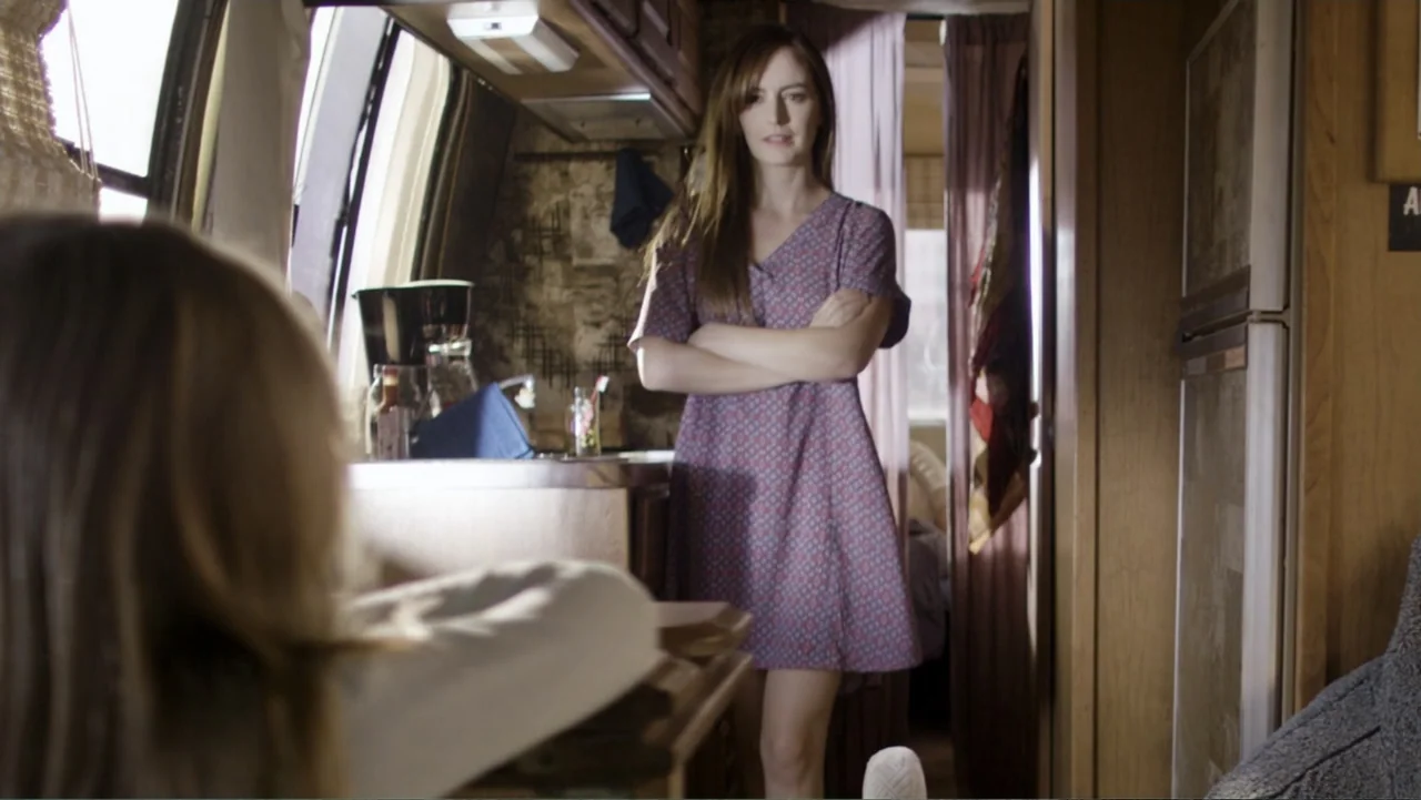 Totem (2022) movie still: medium — The interior of a camper has wood-paneled walls and a small kitchen counter area. A woman stands in the center of…