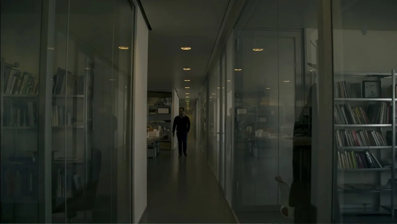 REM (2019) movie still: wide — A long, narrow corridor features glass-walled partitioned spaces lined with metal bookshelves and documents. A man in…