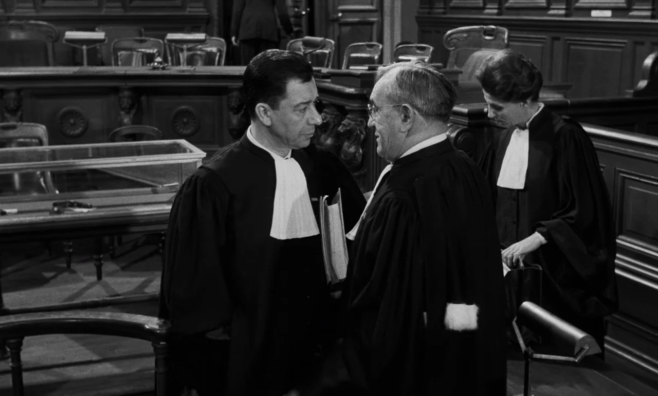 La Verite (1960) movie still: medium — A dark wood-paneled room contains tiered seating and a glass display case. Two men in black robes face each…