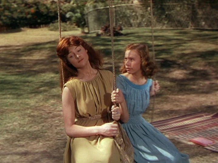 The River (1951) movie still: medium — A grassy lawn features two women sitting on rope swings during the daytime. The area is enclosed by a wire…