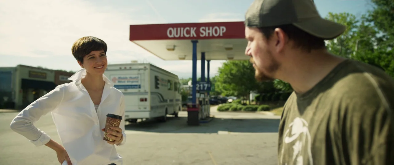 Logan Lucky (2017) movie still: medium — A sunny outdoor gas station forecourt with a red 'Quick Shop' canopy overhead. A woman in a white shirt…