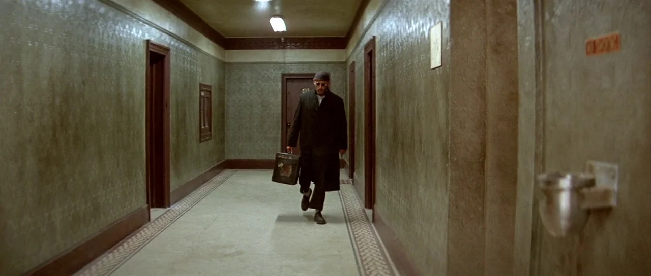 Léon: The Professional movie still: fullbody — A long, narrow apartment building corridor features patterned green wallpaper and multiple dark…