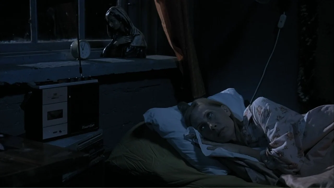 The Man Without a Past (2002) movie still: medium — A dimly lit, cramped bedroom features a window ledge holding a statuette and clock. A woman lies…