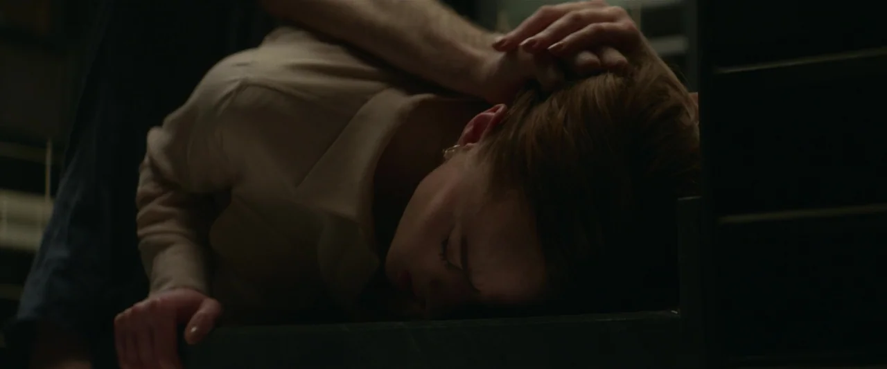 Fair Play (2023) movie still: closeup — A dark, dimly lit indoor staircase features a woman lying flat on a tread. A hand rests gently on the back…