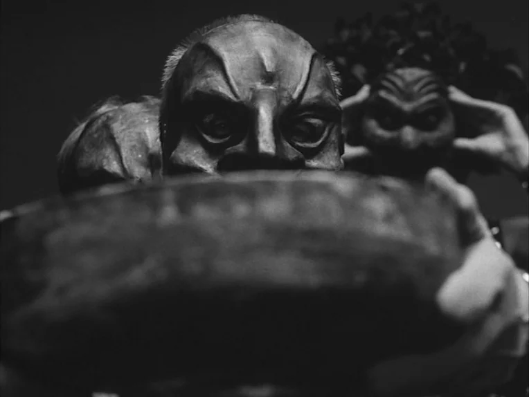 The Rite (1969) movie still: closeup — An indeterminate dark space features a figure wearing a sharp-featured, sculpted theatrical mask. The subject…