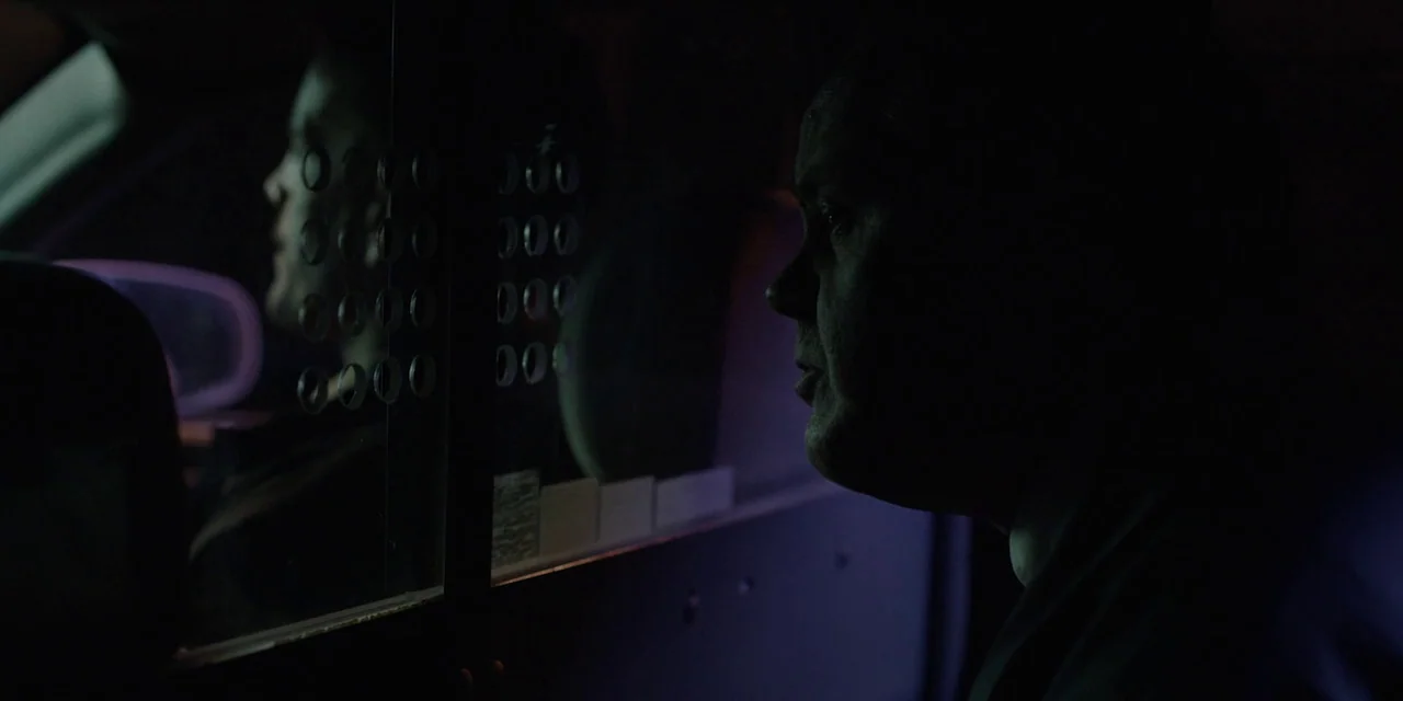 Jakob’s Wife (2021) movie still: medium — The interior of a police vehicle at night features a transparent security partition. Two men are seated on…
