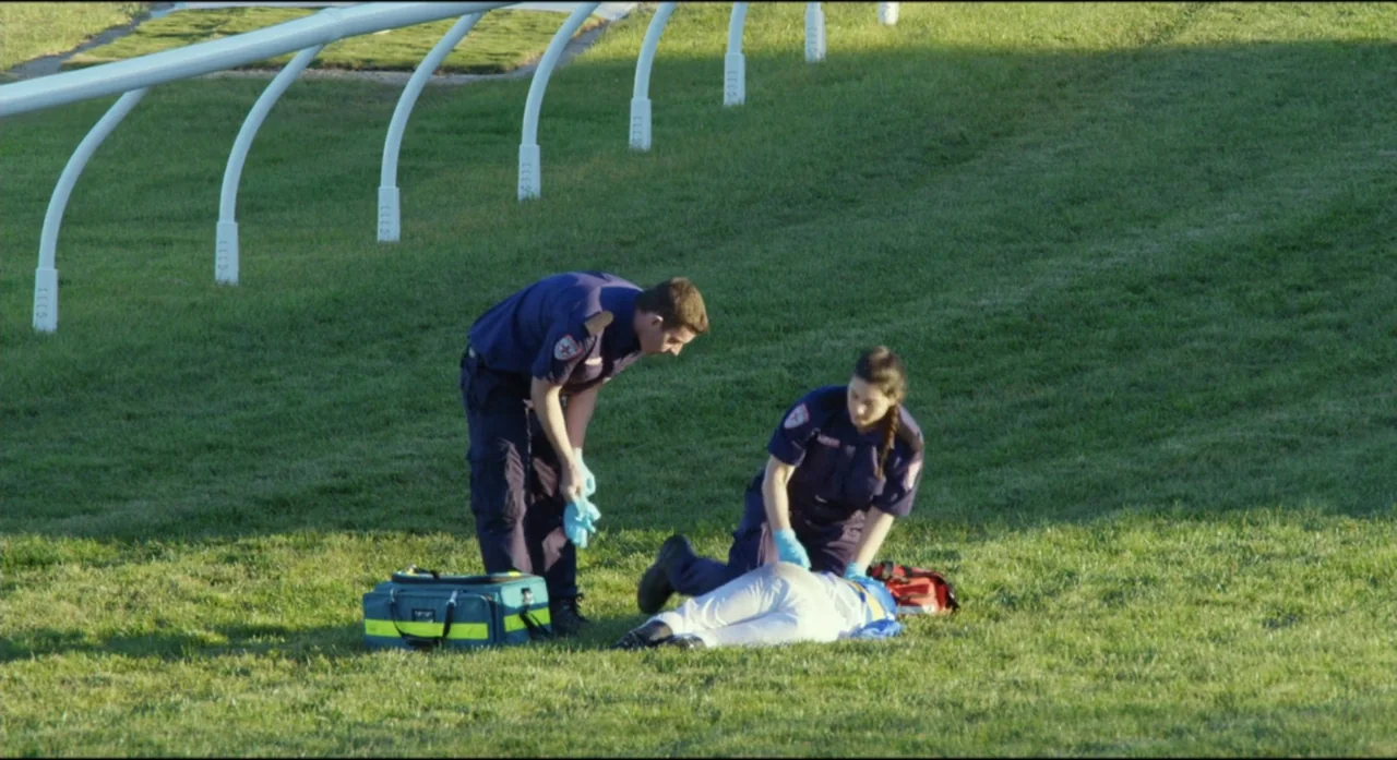 Ride Like a Girl (2019) movie still: wide — A green hillside features a white rail fence bordering the top edge. Two paramedics in navy uniforms…