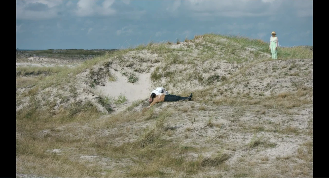 Ninjababy (2021) movie still: wide — A coastal landscape featuring rolling sand dunes covered with patches of wild beach grass. Two people lie on a…