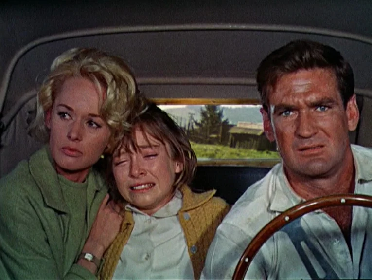 The Birds movie still: medium — The interior of a moving 1960s car viewed from the back seat. A man drives while a woman comforts a young girl who…