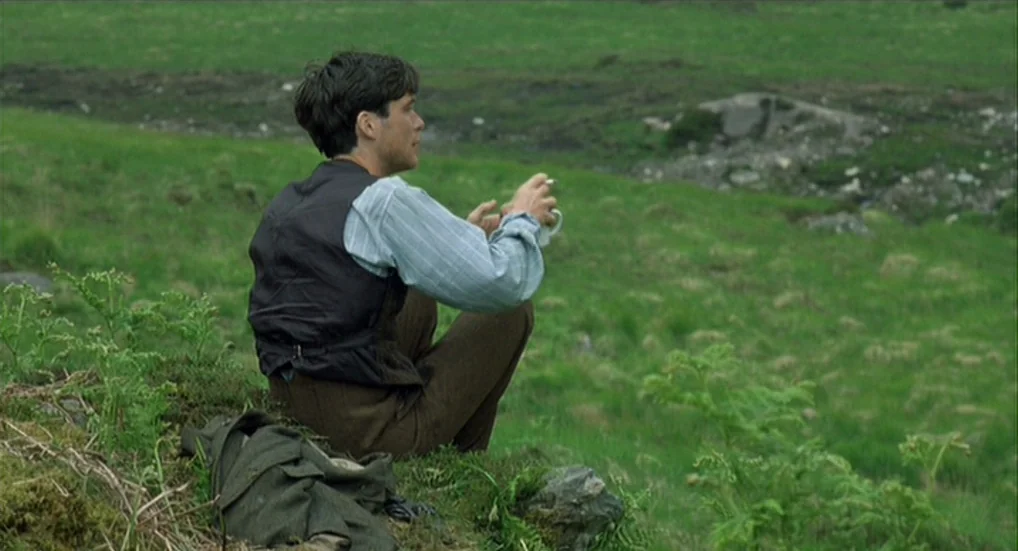 The Wind That Shakes the Barley (2006) movie still: medium — A man sits on a grassy hillside surrounded by an expansive, unpopulated green…