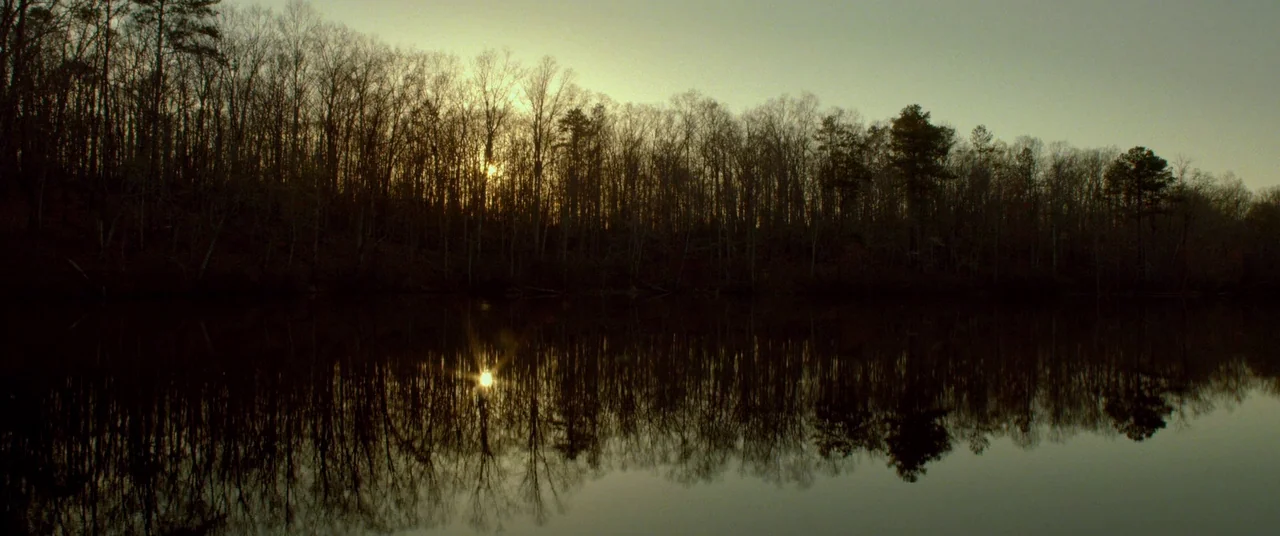 Lawless (2012) movie still: establishing — A dense line of leafless trees borders a quiet lake at sunset. The setting sun reflects clearly in the…