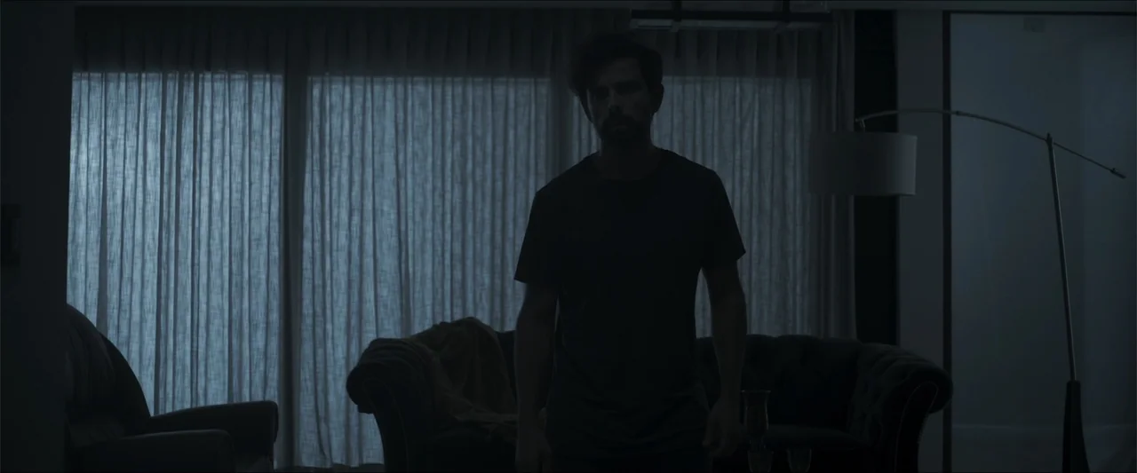 Los habitantes (2023) movie still: medium — A dark, modern living room features a tufted sofa and sheer curtains over windows. A man stands silently…