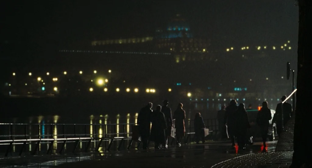 This Night (2008) movie still: wide — A dark river bank at night is illuminated by distant city lights and reflections. A group of individuals in…