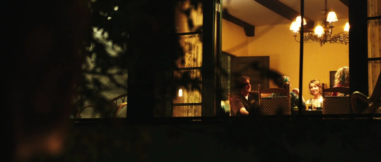 Rampart (2011) movie still: wide — A group of people dine together in a warm, dimly lit residential dining room. The perspective is from outside in…