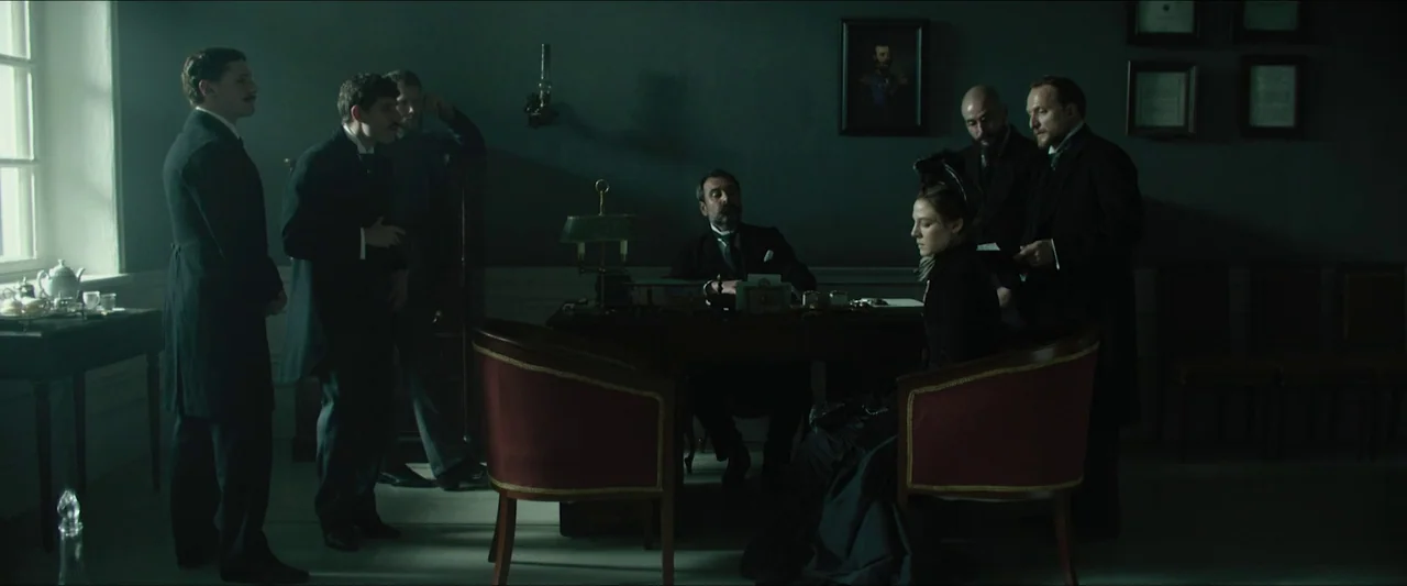 Tchaikovsky’s Wife (2022) movie still: wide — A dimly lit room with dark walls features multiple men standing and sitting around. A woman sits at…