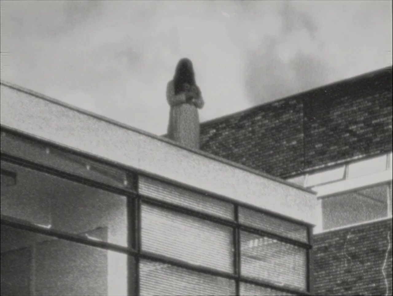 Late Night With The Devil (2023) movie still: wide — A modern building constructed with brick walls and glass facade panels. A lone woman stands on…