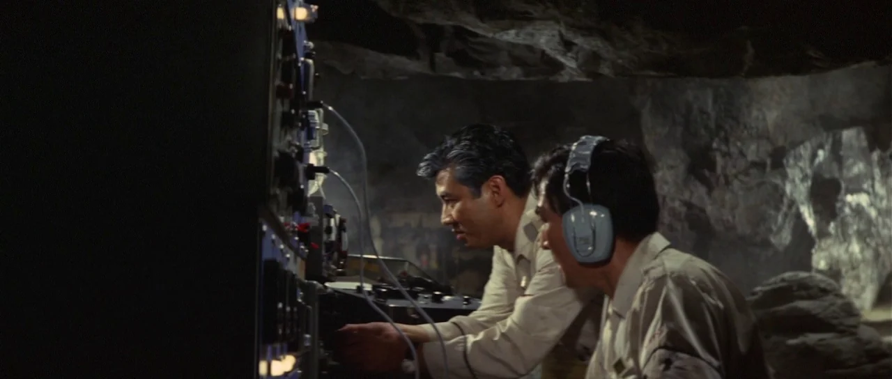 Son of Godzilla (1967) movie still: medium — Two men work at a large electronic console inside a dimly lit stone cave. They operate the equipment…