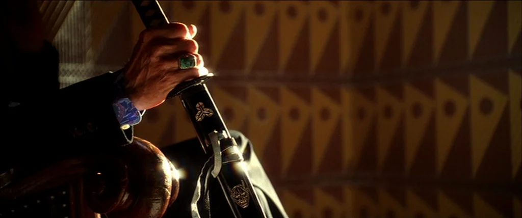 Kill Bill Vol. 1 (2004) movie still: closeup — A close view of an older hand resting on a black lacquer sword hilt. The background features a…
