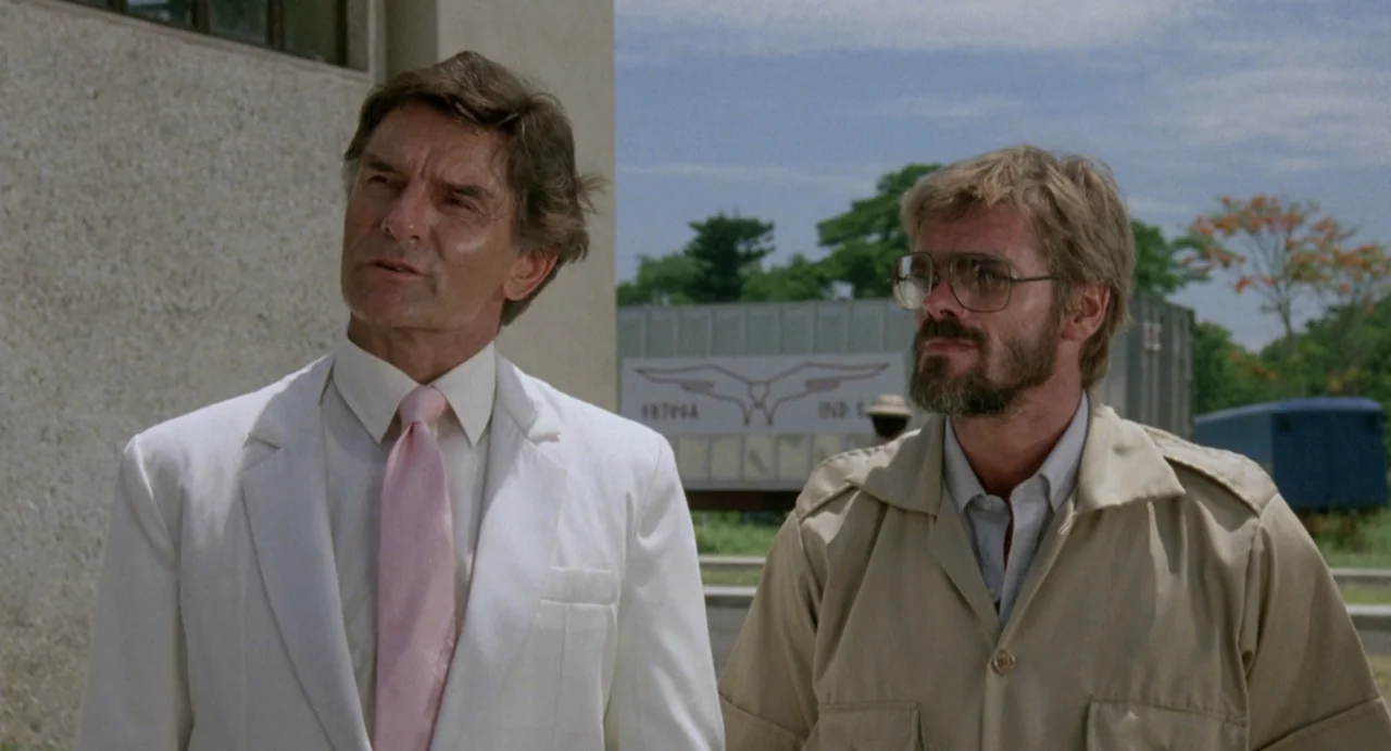 American Ninja (1985) movie still: medium — An outdoor area features industrial buildings with plain beige walls and a trailer visible. Two men…