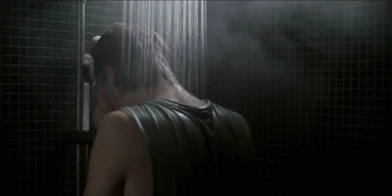 Bring Her Back (2025) movie still: medium — A dark tiled shower stall is filled with steam from an overhead fixture. A young man wearing a wet green…