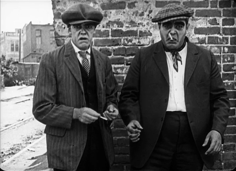 The Kid (1921) movie still: medium — Two men stand against a textured brick wall in an outdoor urban setting. They both wear newsboy caps and suits…