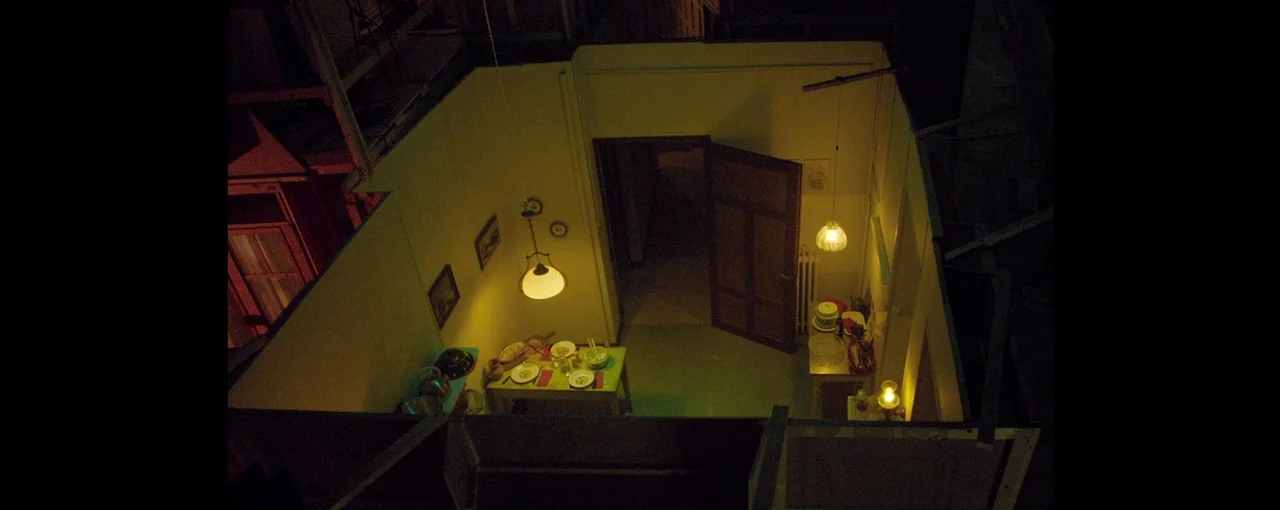 Lux Æterna (2019) movie still: establishing — A top-down view of a small, minimalist interior set featuring a dining area. Two yellow-lit lamps hang…