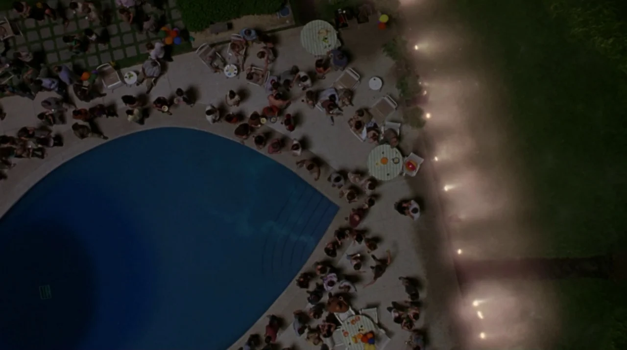 Lady in the Water (2006) movie still: wide — A nighttime aerial view of a crowded poolside patio with round tables and people. Small illuminated…
