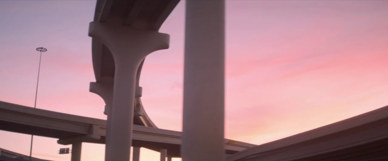 The Unknown Country (2022) movie still: establishing — A complex concrete highway overpass stands against a clear, vibrant pink sunset sky. The…