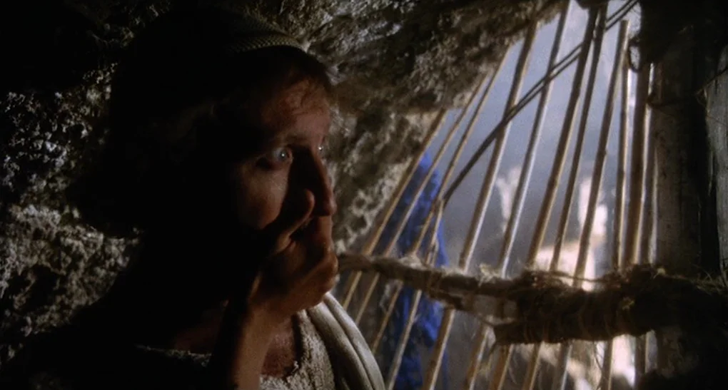 Life of Brian (1979) movie still: closeup — A dimly lit rocky cave interior features rough, jagged stone walls and wooden supports. A man in period…