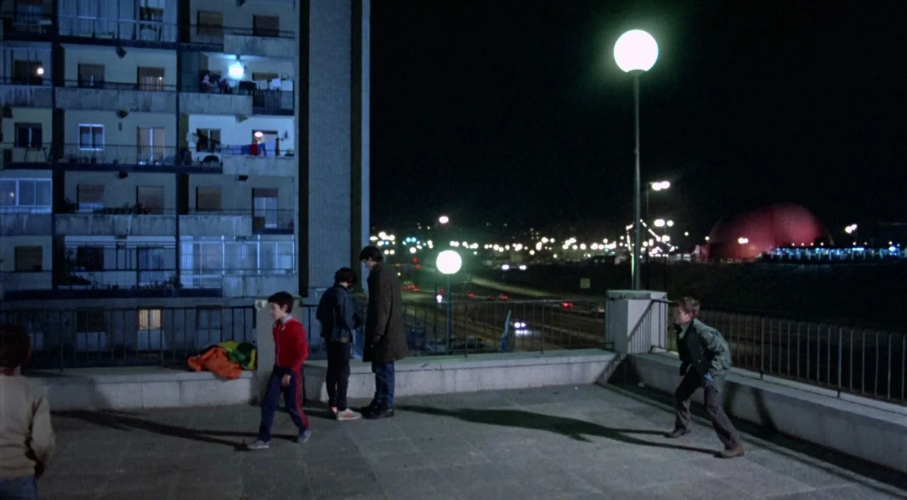 What Have I Done To Deserve This? (1984) movie still: wide — A concrete terrace outside a multi-story apartment block looks out over a dark road. A…