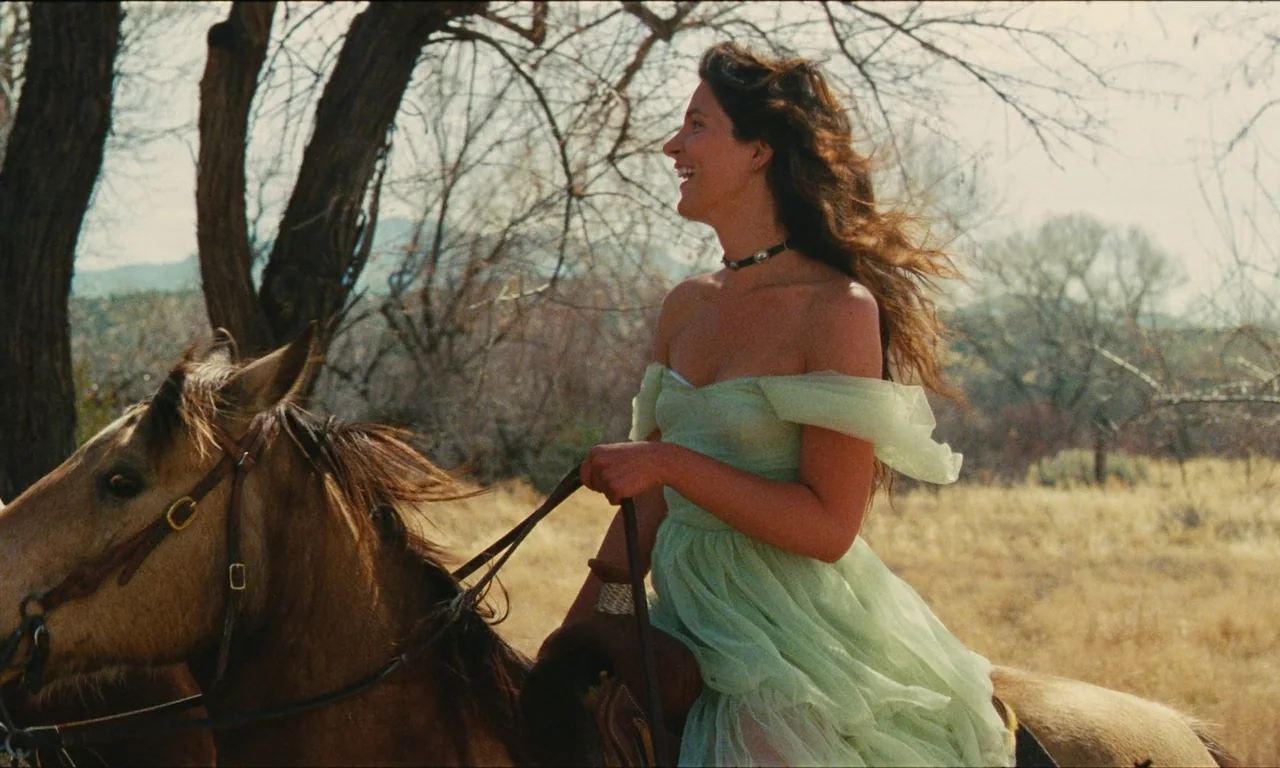 National Anthem (2023) movie still: medium — A woman in a pale green dress rides a brown horse through dry grasslands. Bare trees are visible in the…