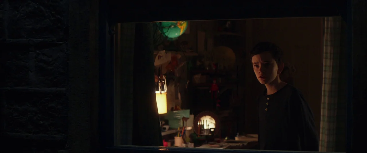 A Monster Calls (2016) movie still: medium — A dark view through a window pane into a cluttered room at night. A boy with short dark hair stands…