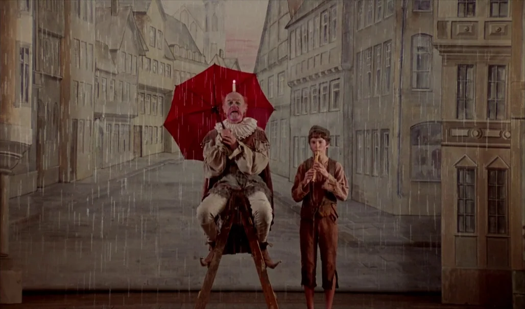 Fanny and Alexander (1982) movie still: fullbody — A theatrical stage set features a painted backdrop of a European medieval city street. A clown…