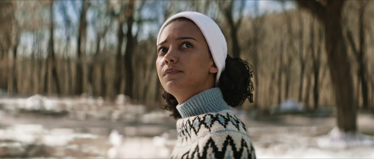 The Sacrifice Game (2023) movie still: medium — A woman stands in a leafless forest during the winter with snow on the ground. She wears a patterned…