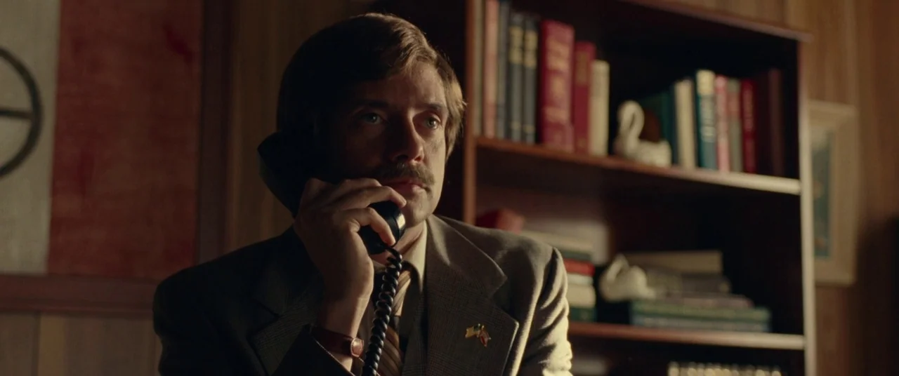 BlacKkKlansman (2018) movie still: medium — A wood-paneled room with a dark bookcase holding various books and decorative swan statues. A man in a…