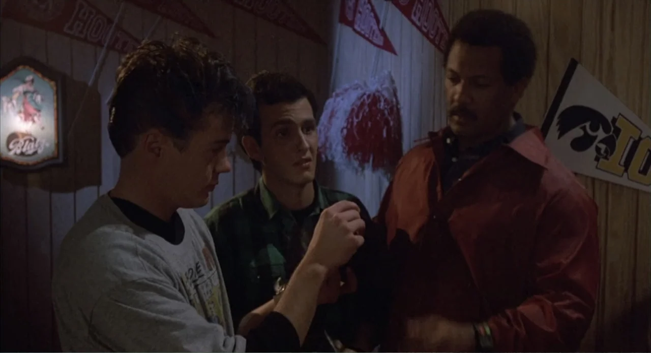 Back to School (1986) movie still: medium — A wood-paneled room features athletic pennants and a lit decorative beer sign on walls. Three men stand…
