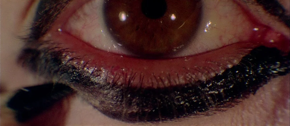 Deep Red (1975) movie still: closeup — A extreme macro view shows a single human eye with thick black makeup applied. A brush or pencil is faintly…