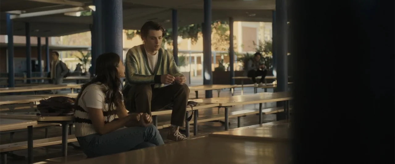Afraid (2024) movie still: medium — A school lunch area features rows of metal benches and blue painted support pillars. Two teenagers are seated…