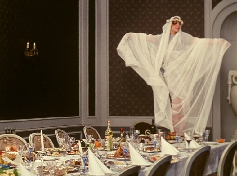 Daisies (1966) movie still: wide — A formal dining room features dark patterned wallpaper and a heavily laden table. A woman stands upon the table…