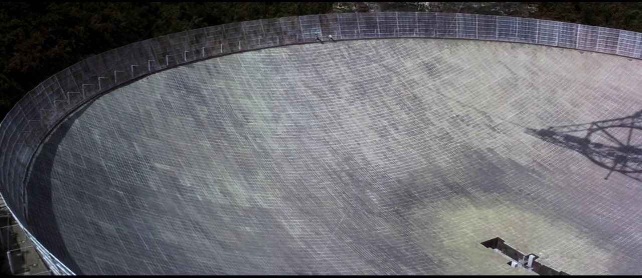 GoldenEye (1995) movie still: wide — A massive, circular radio telescope dish sits nestled in a dense, green jungle setting. Two tiny figures sprint…