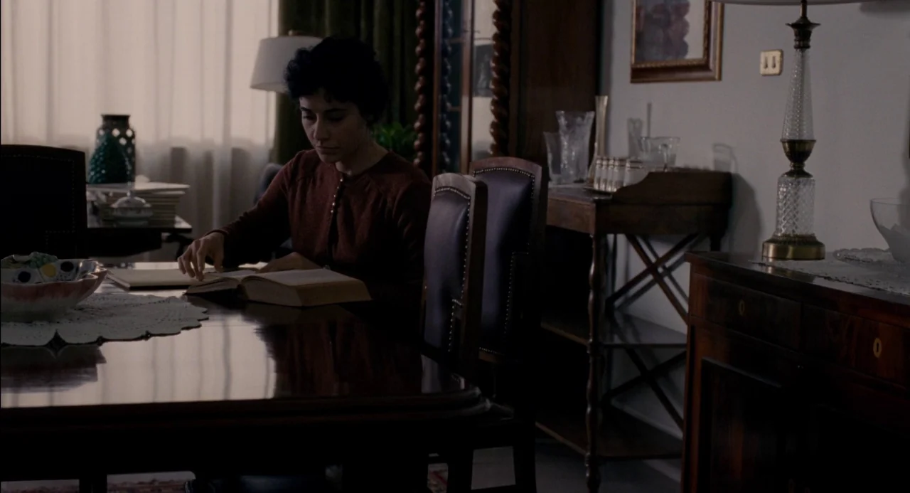Pasolini (2014) movie still: medium — A quiet room featuring a dark polished wooden table and vintage-style furniture pieces. A person sits alone,…