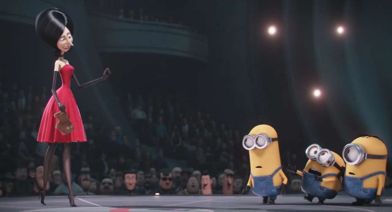 Minions (2015) movie still: fullbody — An auditorium stage features a tall woman in a red dress addressing three yellow creatures. A darkened crowd…