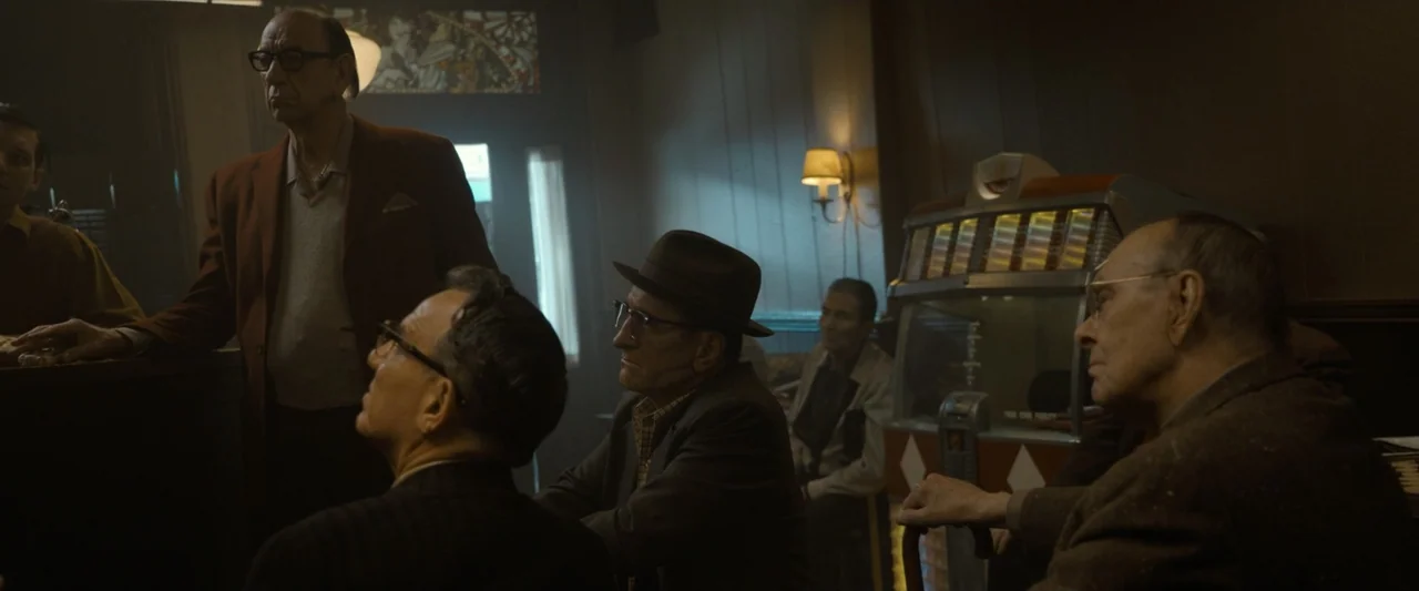 The Alto Knights (2025) movie still: medium — A dark, wood-paneled interior of a 1950s establishment featuring a vintage jukebox. Several older men…