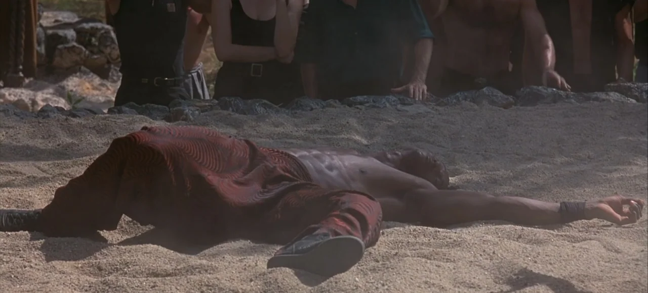 Mortal Kombat (2021) movie still: medium — A sand pit surrounded by a stone border with a group of onlookers standing above. A muscular man lies…