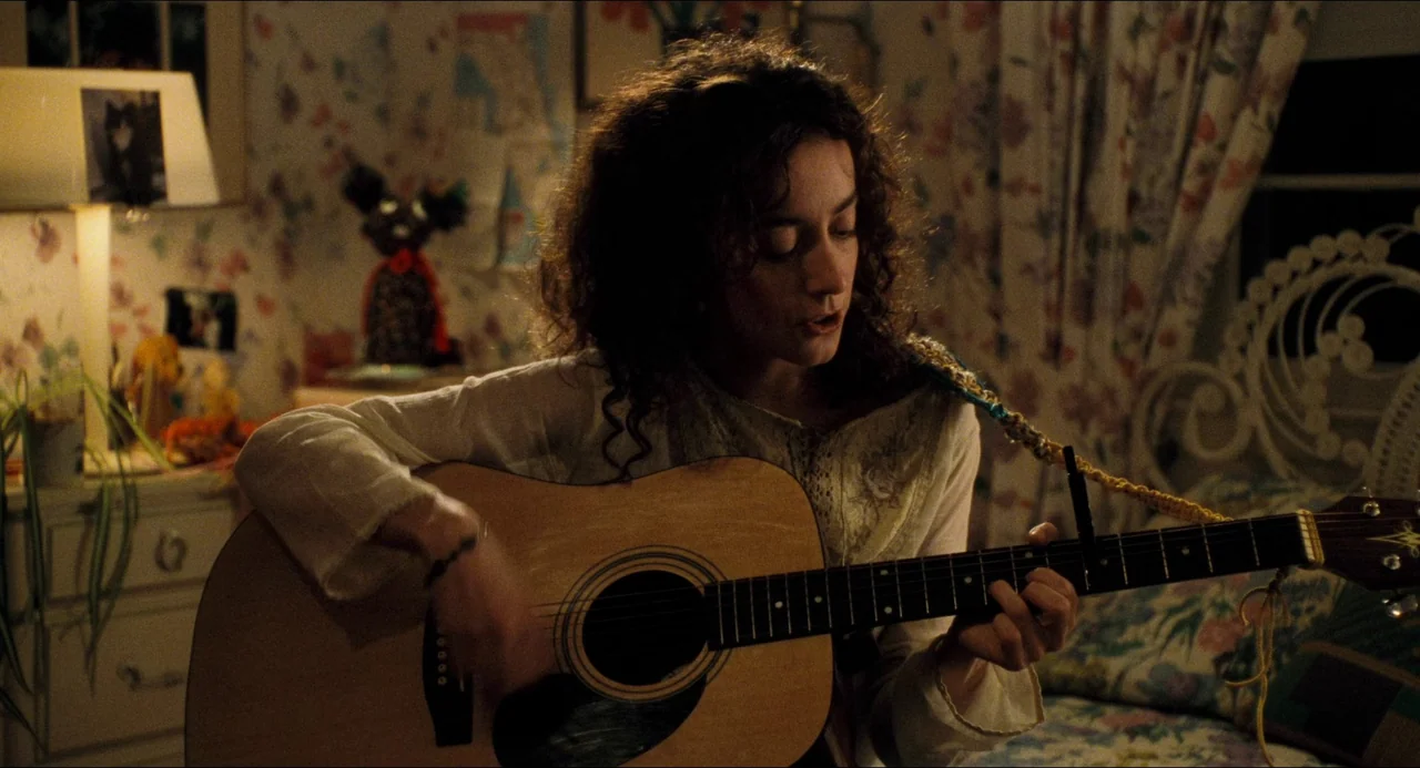 Happiness (1998) movie still: medium — A dimly lit room with floral wallpaper and a lamp in the background. A woman with curly hair plays an…